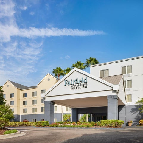 A Fairfield Inn & Suites building features a covered entrance with landscaping and a multi-story white facade under a blue sky.