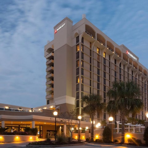 A Marriott hotel building with a light-colored facade, palm trees, and outdoor lighting at dusk.