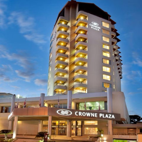 A tall, multi-story Crowne Plaza hotel building is shown with balconies and illuminated windows against a twilight sky.