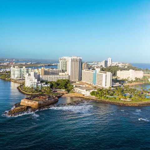 An aerial view showcases a large resort complex with multiple buildings, a peninsula, and the ocean surrounding it.