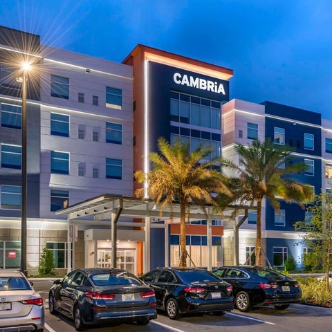 A modern Cambria hotel building with palm trees, a covered entrance, and several cars parked in front.