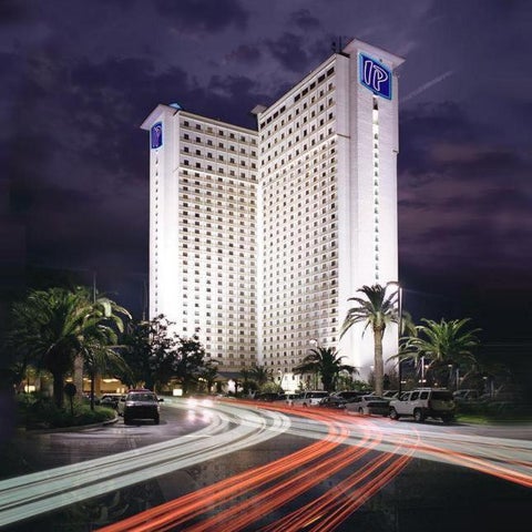 A tall, white hotel building with a prominent 'IP' logo is illuminated at night, surrounded by palm trees and moving traffic.