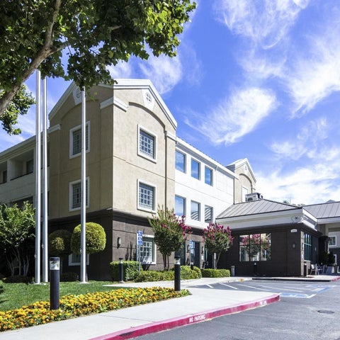 A large, multi-story hotel building with a light-colored facade, landscaping, and a covered entryway under a bright sky.