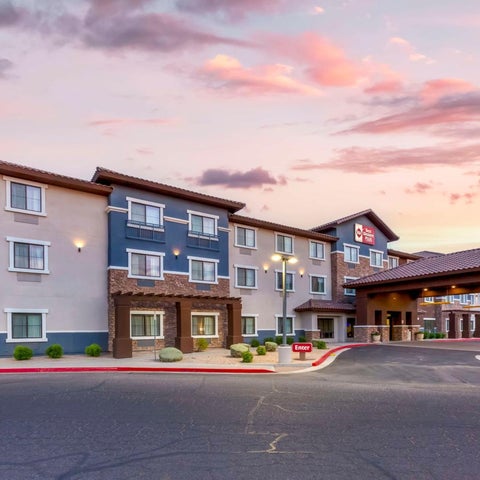A multi-story hotel with a blue and beige facade sits under a colorful sunset sky with a covered entryway and parking lot.