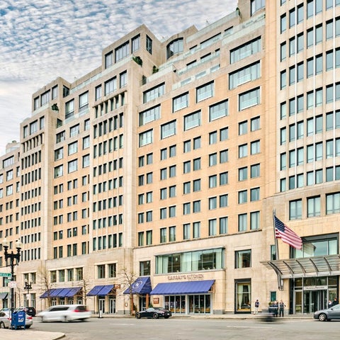 A large, modern building with a light-colored facade features a street-level entrance with flags and shops.
