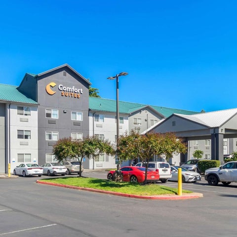 A Comfort Suites hotel is shown with a parking lot, trees, and several vehicles parked in front of the building.