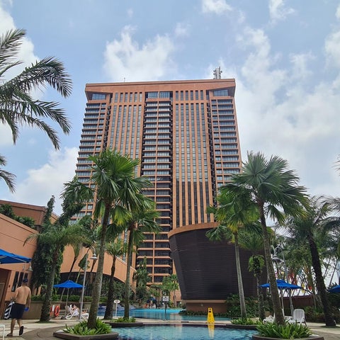 A tall, multi-story building with a pool and palm trees in the foreground, under a partly cloudy sky.