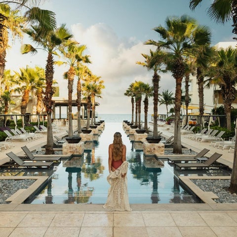 A woman in a long dress stands facing a pool reflecting palm trees and a hotel, with lounge chairs and a beachfront setting.