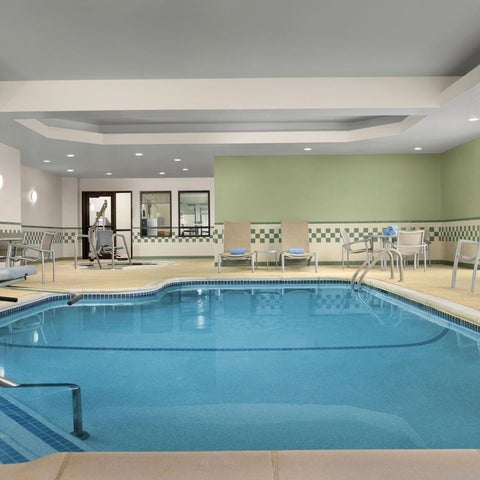 An indoor pool with a light blue hue is surrounded by lounge chairs and tables in a well-lit hotel space.