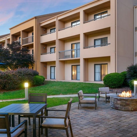 A multi-story hotel building features an outdoor patio with seating, a fire pit, and lush landscaping.