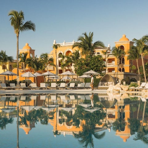 A large, rectangular pool reflects a vibrant, orange-colored hotel building and palm trees under a clear blue sky.