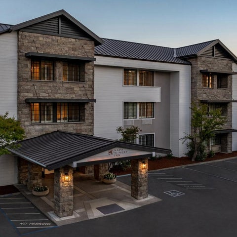 A three-story hotel building with a stone and stucco facade, a covered entrance, and a parking lot in front.