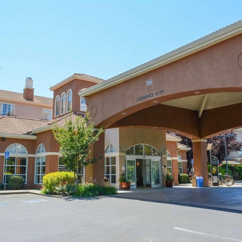 A tan-colored hotel building features a covered entrance with arched windows and landscaping in front.