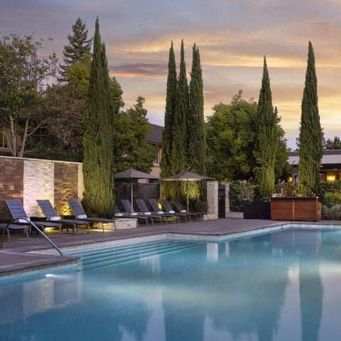 A luxurious outdoor pool area features lounge chairs, umbrellas, and a reflecting pool with tall cypress trees against a sunset sky.