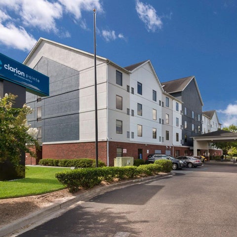 A Clarion Pointe hotel building facade with a parking lot, landscaping, and a blue sign visible under a partly cloudy sky.