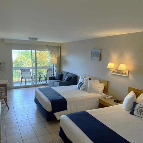 A hotel room features two queen beds, a sofa, and a balcony with outdoor seating.