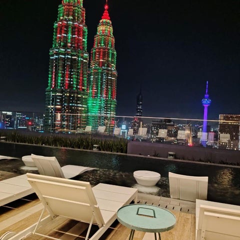 An outdoor pool area with lounge chairs and a table overlooks the illuminated Petronas Twin Towers and Kuala Lumpur skyline at night.