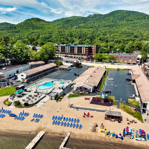 An aerial view shows a lakeside resort with a sandy beach, pool, lounge chairs, and several motel-style buildings.