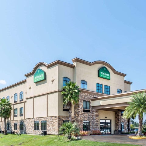 A two-story Wingate hotel with a light-colored facade, palm trees, and a covered entrance sits against a clear blue sky.