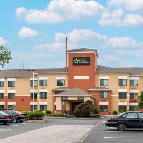 A StayAmerica hotel building with a multi-story facade, parking lot, and trees visible under a cloudy sky.