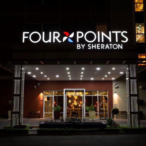 The entrance of a Four Points by Sheraton hotel is illuminated at night, featuring a lit sign and landscaped area.