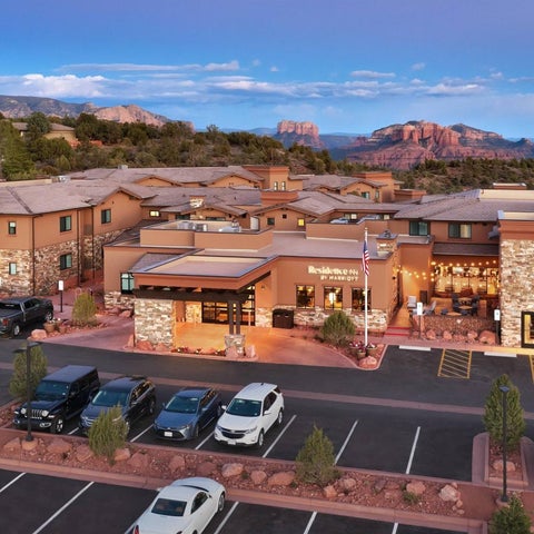 A large, multi-story hotel with a stone facade sits near red rock formations under a partly cloudy sky.