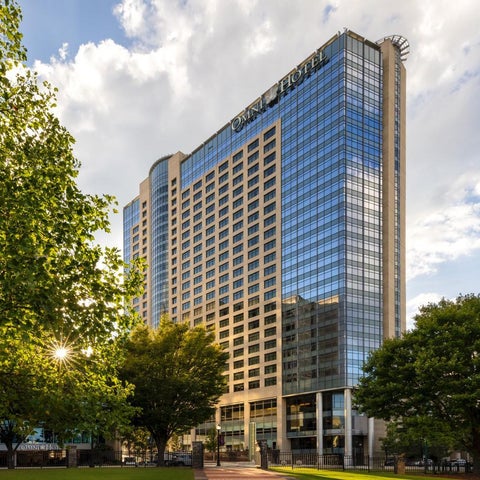 A tall, modern hotel building with a glass facade is framed by trees and a partly cloudy sky.
