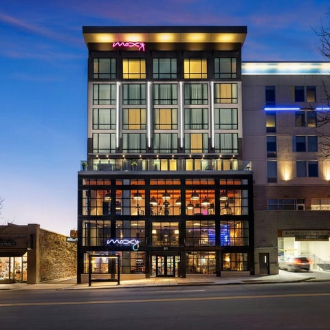 A modern hotel building with a neon sign and glass-enclosed lobby is shown at dusk.