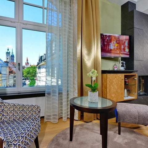 A hotel room features a patterned armchair, a small table with ottoman, and a view of a cityscape through sheer curtains.
