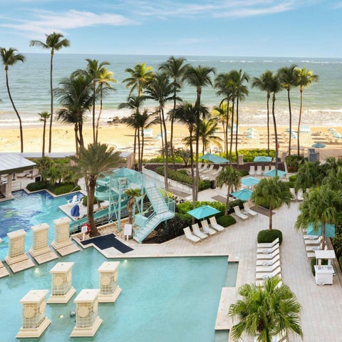 A resort features a large pool with water slides and lounge chairs overlooking a sandy beach and palm trees.