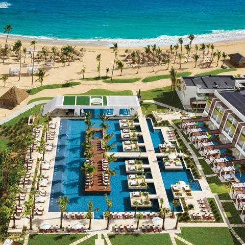 An aerial view showcases a large resort with a sprawling pool complex, beach access, and multiple buildings with lounge areas.