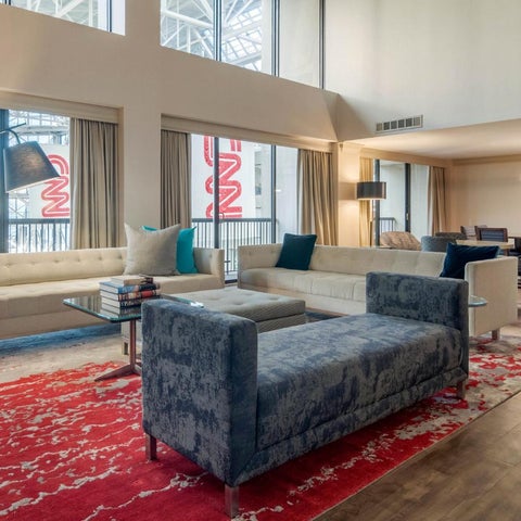 A modern living room features a sectional sofa, accent chairs, and a patterned rug against a backdrop of large windows.
