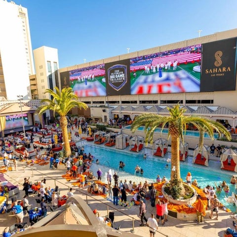 A large pool area at the Sahara Las Vegas hotel features a vibrant crowd, palm trees, and a large digital display.