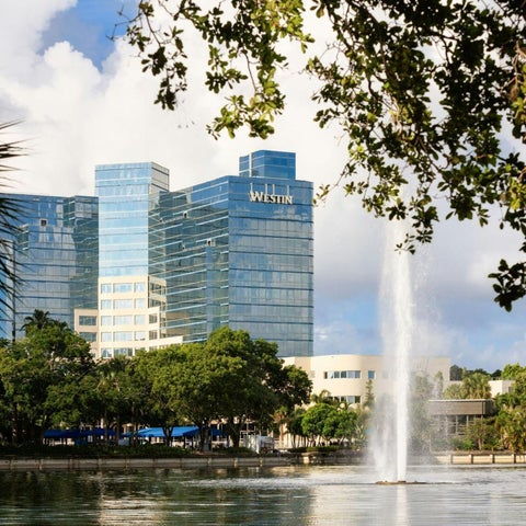 A modern Westin hotel building is reflected in a pond with a fountain and lush greenery in the foreground.