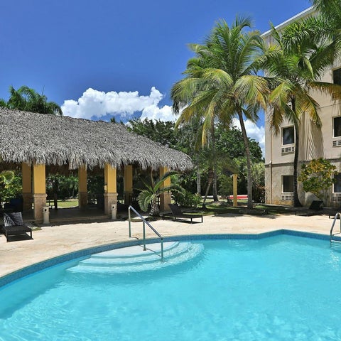 A turquoise pool with lounge chairs and a thatched-roof cabana sits in front of a multi-story building with palm trees.