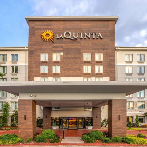 A La Quinta Inn & Suites building features a tan facade, prominent logo, covered entrance, and lush landscaping.