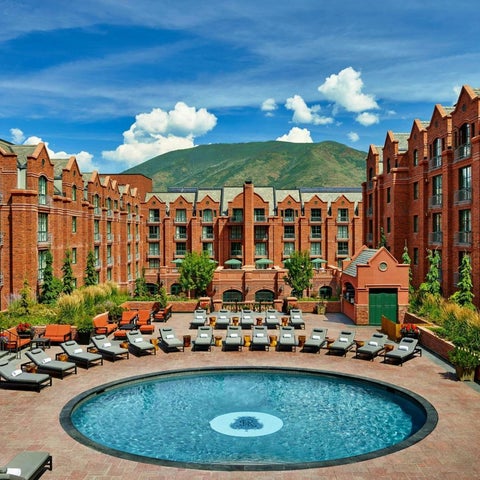 A large circular pool with lounge chairs surrounds a courtyard area in front of a brick building with a mountain backdrop.