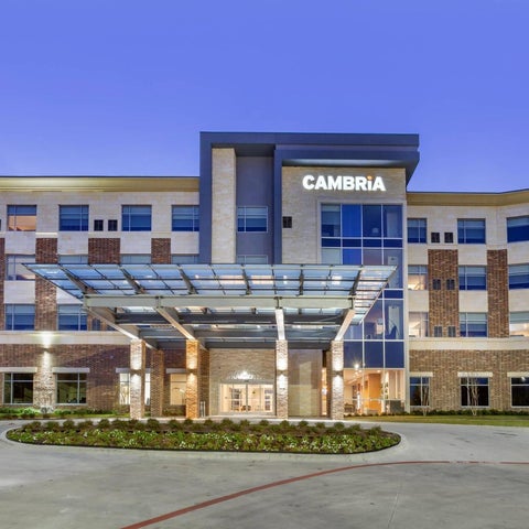 A modern Cambria hotel building features a brick and glass facade with a covered entrance and well-maintained landscaping.