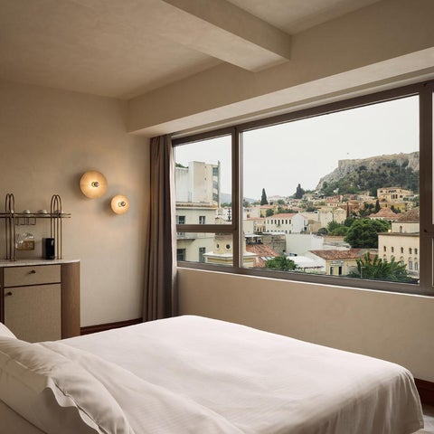 A hotel room features a white bed, a desk with a chair, and a large window overlooking a cityscape with a historic landmark.