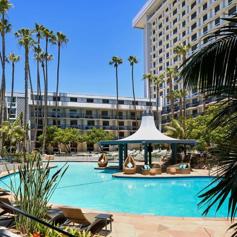 A bright blue pool with lounge chairs and a shaded cabana is surrounded by palm trees and a large hotel building.