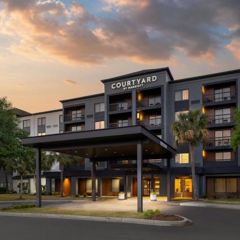 A Courtyard by Marriott hotel building is shown with a covered entrance and lush greenery under a cloudy sunset sky.