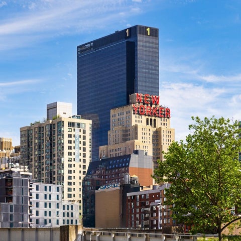 The New Yorker hotel rises above a cluster of buildings in a dense New York City skyline under a partly cloudy sky.