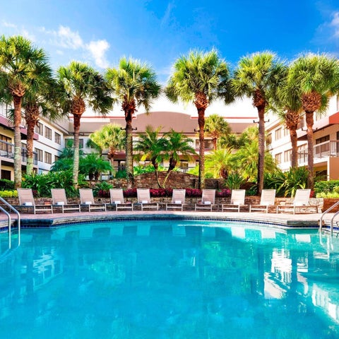 A bright blue pool with lounge chairs and palm trees is surrounded by a hotel building with balconies.