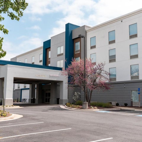 A modern hotel building with a covered entrance and parking lot, featuring a light-colored facade and blue accents.