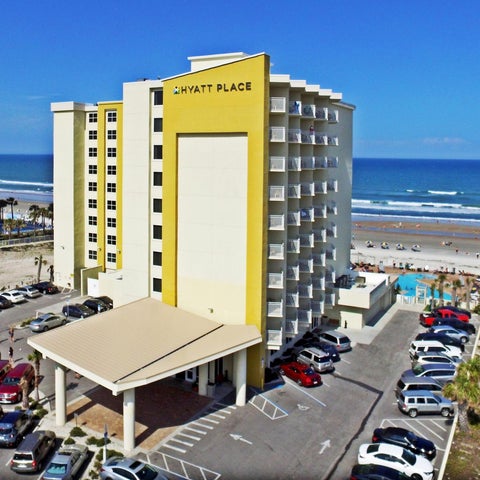 A Hyatt Place hotel building is situated near a beach with a parking lot and pool area visible in the foreground.