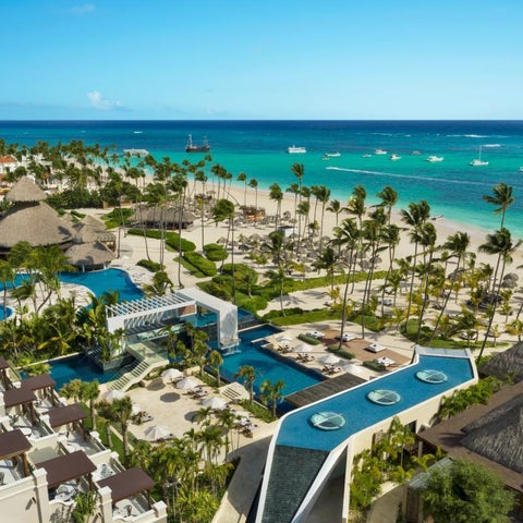 An aerial view showcases a luxury resort with multiple pools, a sandy beach, palm trees, and turquoise ocean waters.