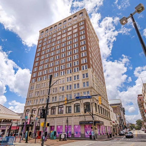 A tall, brick building with multiple stories stands on a city street with traffic lights and signage.