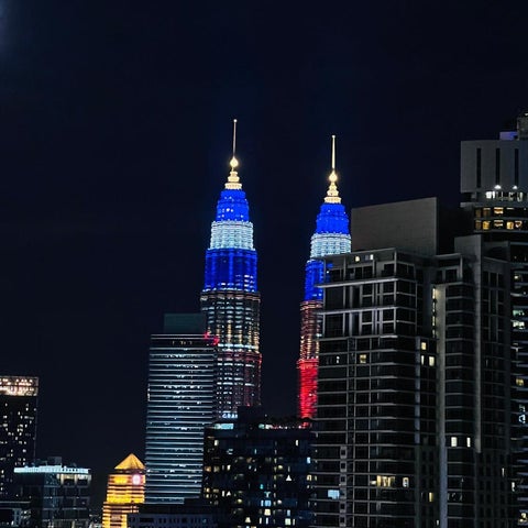 The Petronas Twin Towers are illuminated in blue and red against a dark night sky, surrounded by other city buildings.