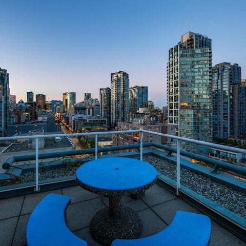 A balcony with a small blue table and two chairs overlooks a cityscape of modern high-rise buildings at dusk.