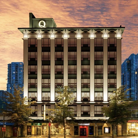 A modern building with a large 'Q' logo on top, featuring a symmetrical facade and surrounding trees and shops.